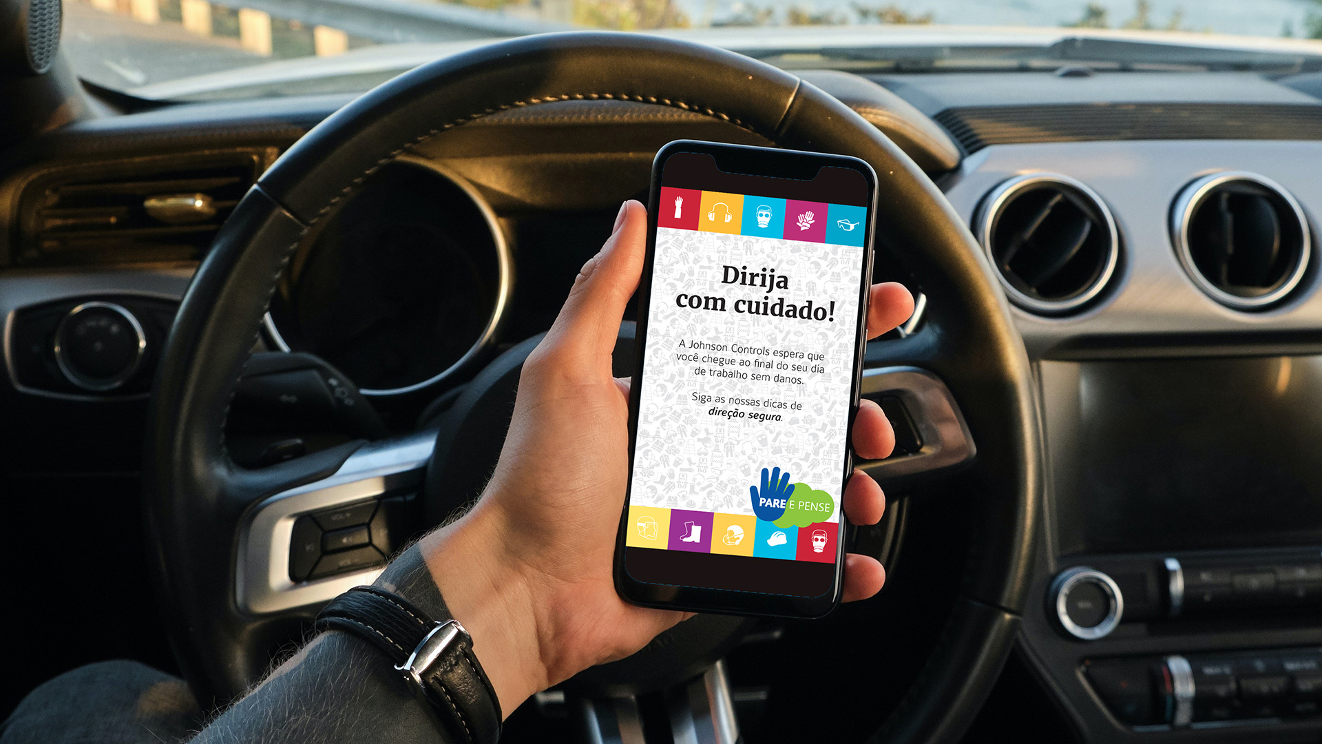 Imagem de mão de homem segurando um celular dentro do carro com a comunicação da campanha de endomarketing Pare & Pense Johnson Controls na tela.