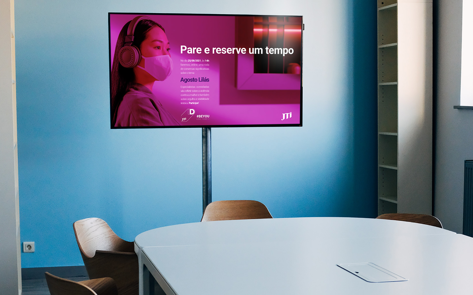 Imagem de sala de reunião com TV e imagem da campanha Agosto Lilás JTI na tela.