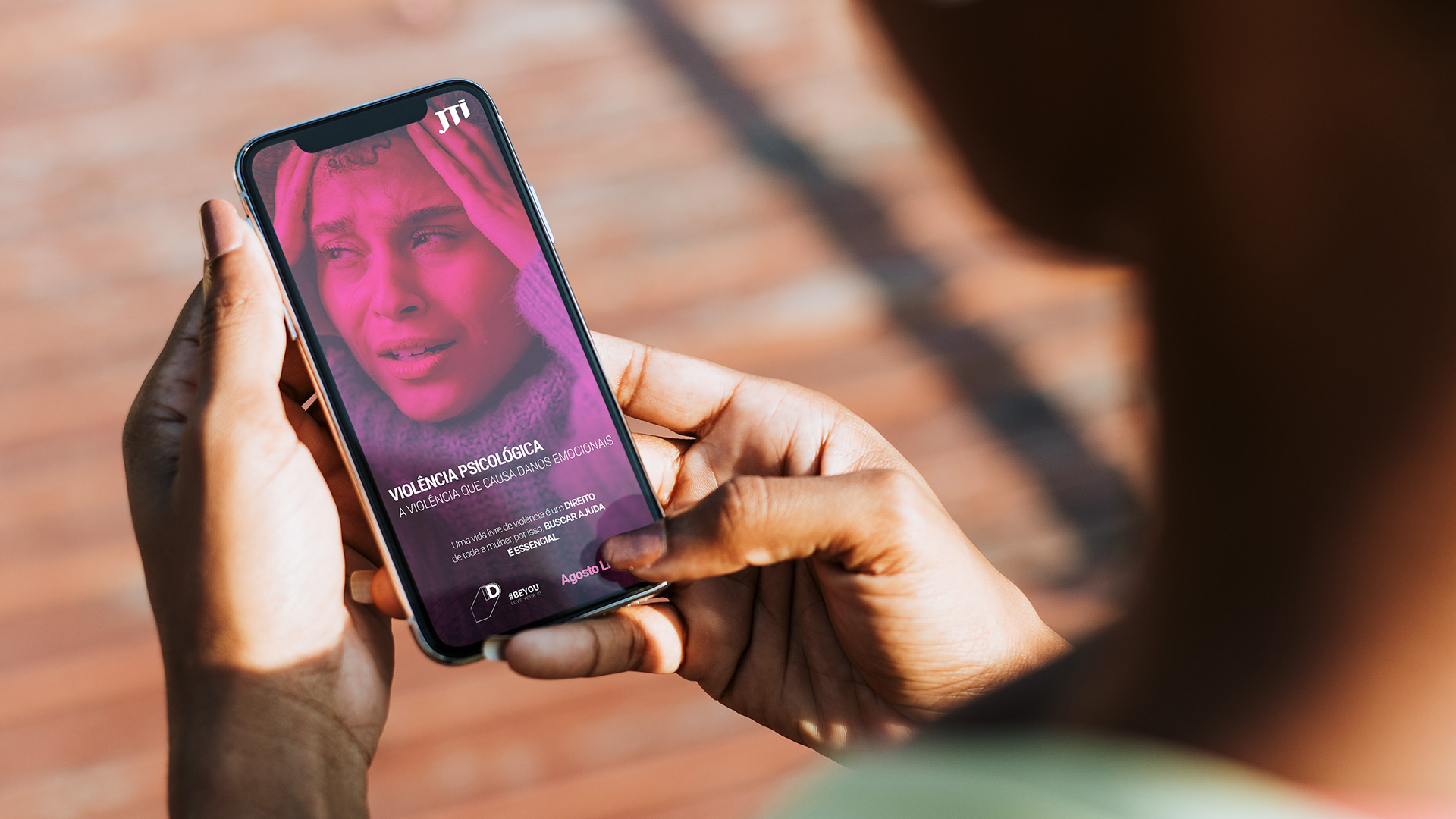 Imagem de smartphone na mão de uma pessoa com imagem da campanha Agosto Lilás JTI na tela.